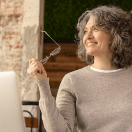 Na imagem, uma mulher sorrindo está em um café. Ela está sentada perto de uma mesa com um notebook e em sua mão esquerda segura um óculos.