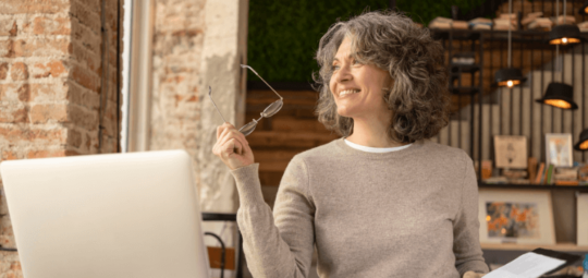 Na imagem, uma mulher sorrindo está em um café. Ela está sentada perto de uma mesa com um notebook e em sua mão esquerda segura um óculos.