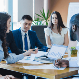 Na imagem, duas mulheres e dois homens estão sentados ao redor de uma mesa de trabalho. Nessa mesa há um notebook com um gráfico na tela.