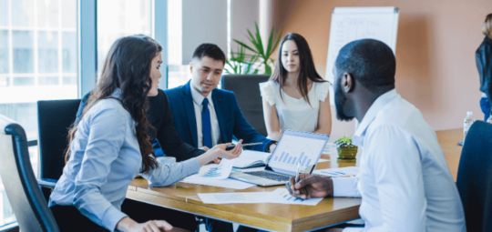 Na imagem, duas mulheres e dois homens estão sentados ao redor de uma mesa de trabalho. Nessa mesa há um notebook com um gráfico na tela.