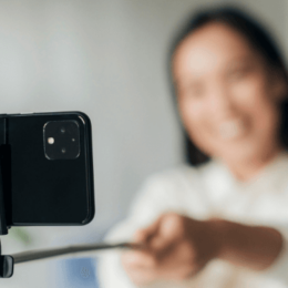 Na foto, uma mulher sorridente posa para uma selfie com um smartphone preso a um bastão para foto.