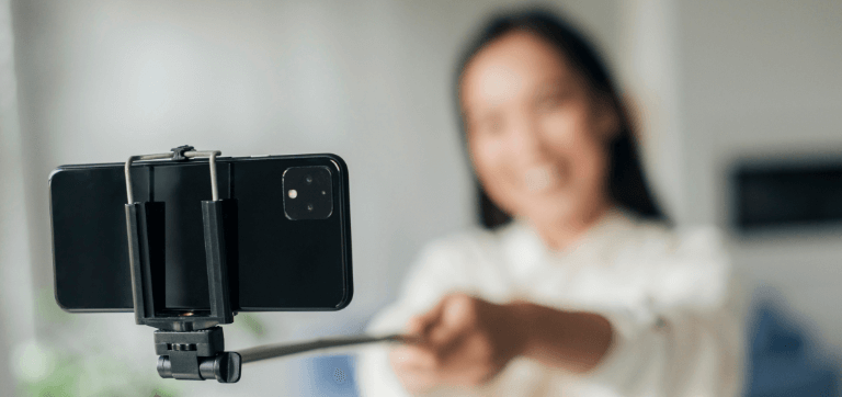 Na foto, uma mulher sorridente posa para uma selfie com um smartphone preso a um bastão para foto.