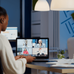 Na imagem, uma mulher está sentada perto de uma mesa de trabalho. Na mesa está um notebook com imagens de pessoas na tela, representando uma chamada de vídeo virtual de trabalho.