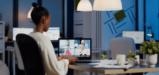 Na imagem, uma mulher está sentada perto de uma mesa de trabalho. Na mesa está um notebook com imagens de pessoas na tela, representando uma chamada de vídeo virtual de trabalho.