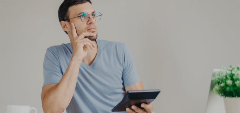 Na imagem está um homem sentado ao redor de uma mesa. Ele está pensativo, com uma mão no queixo e a outra segurando uma calculadora.