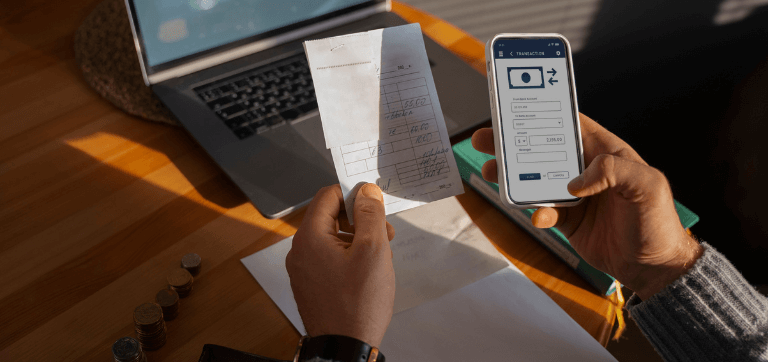 No foco central da imagem mostra uma pessoa segurando uma folha com valores enquanto na outra mão segura o celular para fazer uma transferência. Como plano de fundo está uma mesa de madeira, um notebook, um livro e algumas moedas dispostos sobre ela.