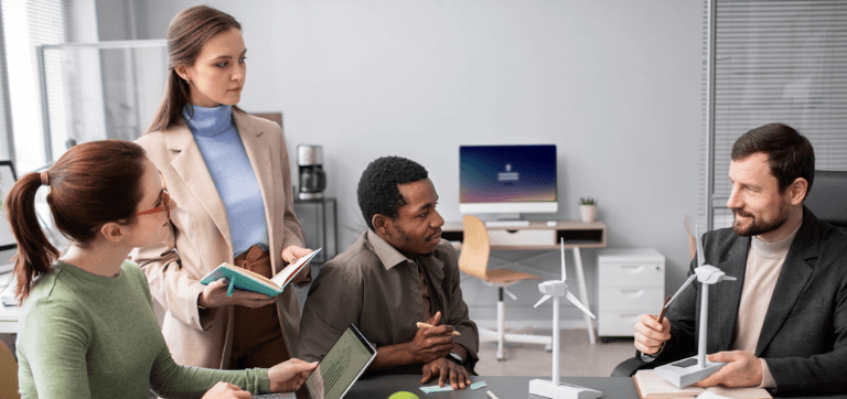 Na imagem, dois homens e duas mulheres estão ao redor de uma mesa de trabalho conversando. Um deles segura um mini gerador de energia eólica, outro uma caneta, uma mulher segura um bloco de notas e a outra um notebook.