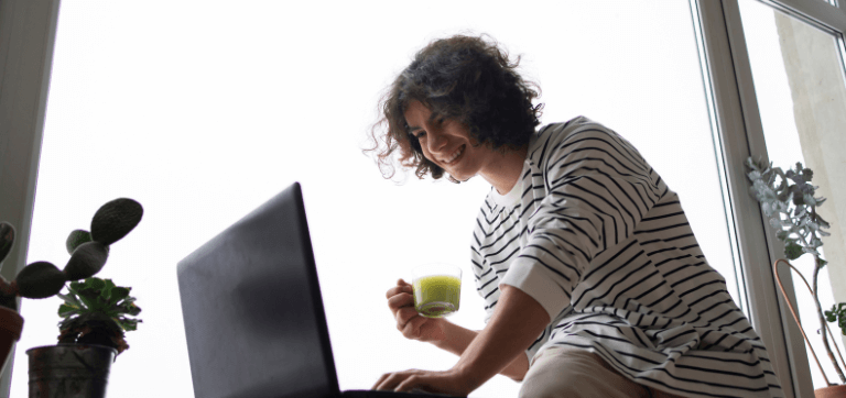 Na imagem está um homem jovem sentado na frente de uma grande janela de vidro. À sua frente está um notebook, que ele utiliza com uma mão enquanto segura uma xícara com a outra.