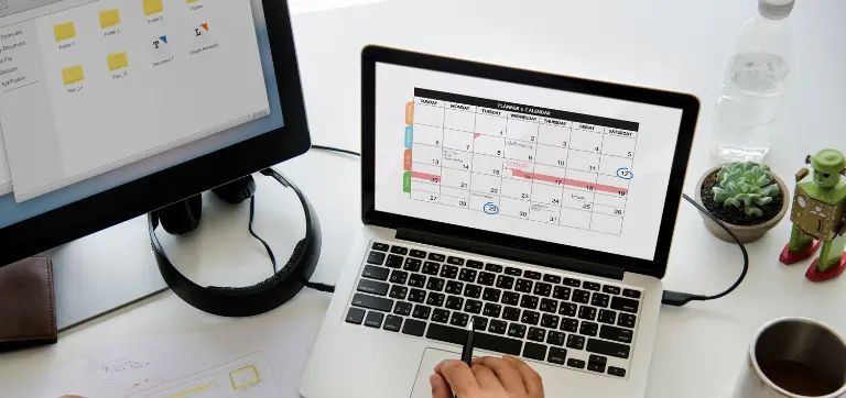 Una persona trabajando en una laptop que muestra un calendario, con un segundo monitor, auriculares y suministros de oficina cerca.