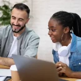 Dos personas en una mesa con laptops, una sosteniendo un teléfono inteligente, en un entorno de oficina moderna.