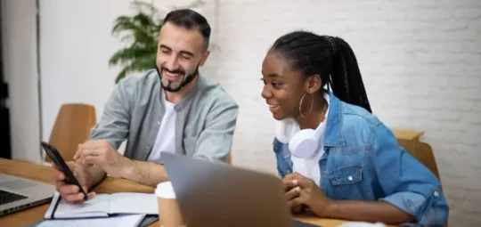 Dos personas en una mesa con laptops, una sosteniendo un teléfono inteligente, en un entorno de oficina moderna.