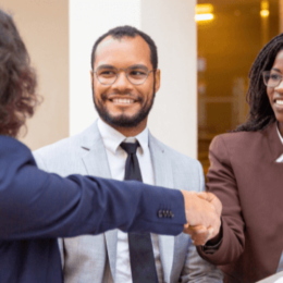 Na imagem, três pessoas estão em uma reunião de negócios. Duas mulheres se cumprimentam e um homem presencia o momento.