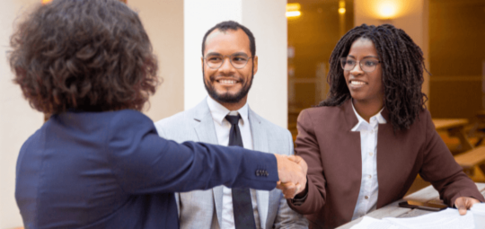 Na imagem, três pessoas estão em uma reunião de negócios. Duas mulheres se cumprimentam e um homem presencia o momento.