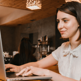 Na imagem, uma mulher está em um café trabalhando. Ela sorri para a tela do computador enquanto digita.