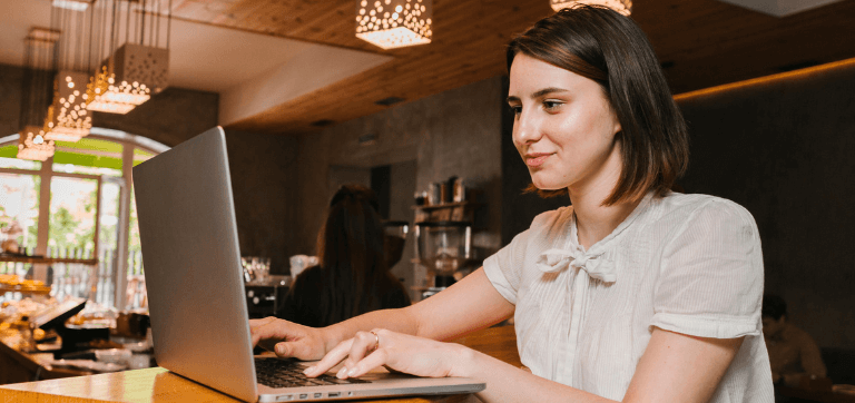 Na imagem, uma mulher está em um café trabalhando. Ela sorri para a tela do computador enquanto digita.