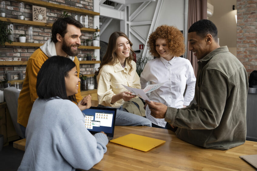 Como centro da imagem, pessoas estão trabalhando juntas e em reunião. Como plano de fundo há um ambiente corporativo, com plantas, mesa e paredes.