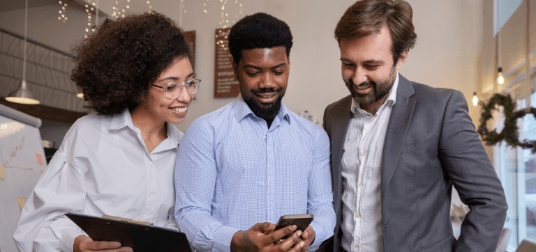 Na imagem, há três pessoas em um ambiente corporativo. Há um homem que segura um celular no centro, ao lado dele está uma mulher que olha para o celular sorridente e outro homem ao lado deles também sorri.