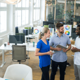 Na imagem há três pessoas conversando. São dois homens e uma mulher em um espaço corporativo, com computadores e mesas.