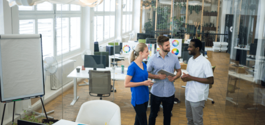 Na imagem há três pessoas conversando. São dois homens e uma mulher em um espaço corporativo, com computadores e mesas.