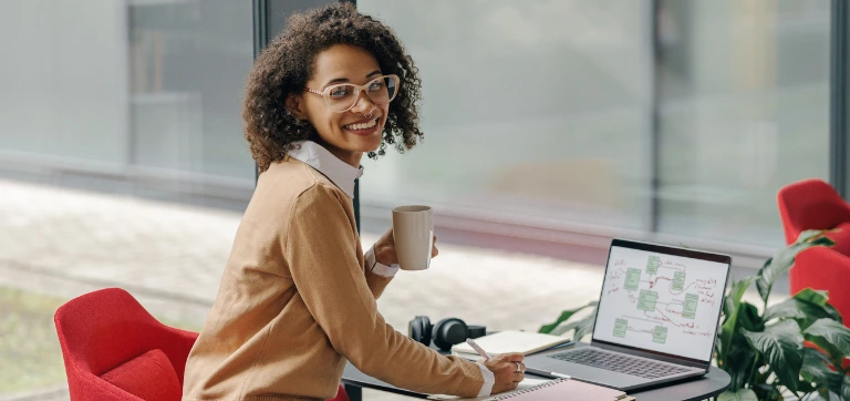 Persona en un escritorio con computadora portátil, taza de café en mano, en un entorno de oficina moderno.